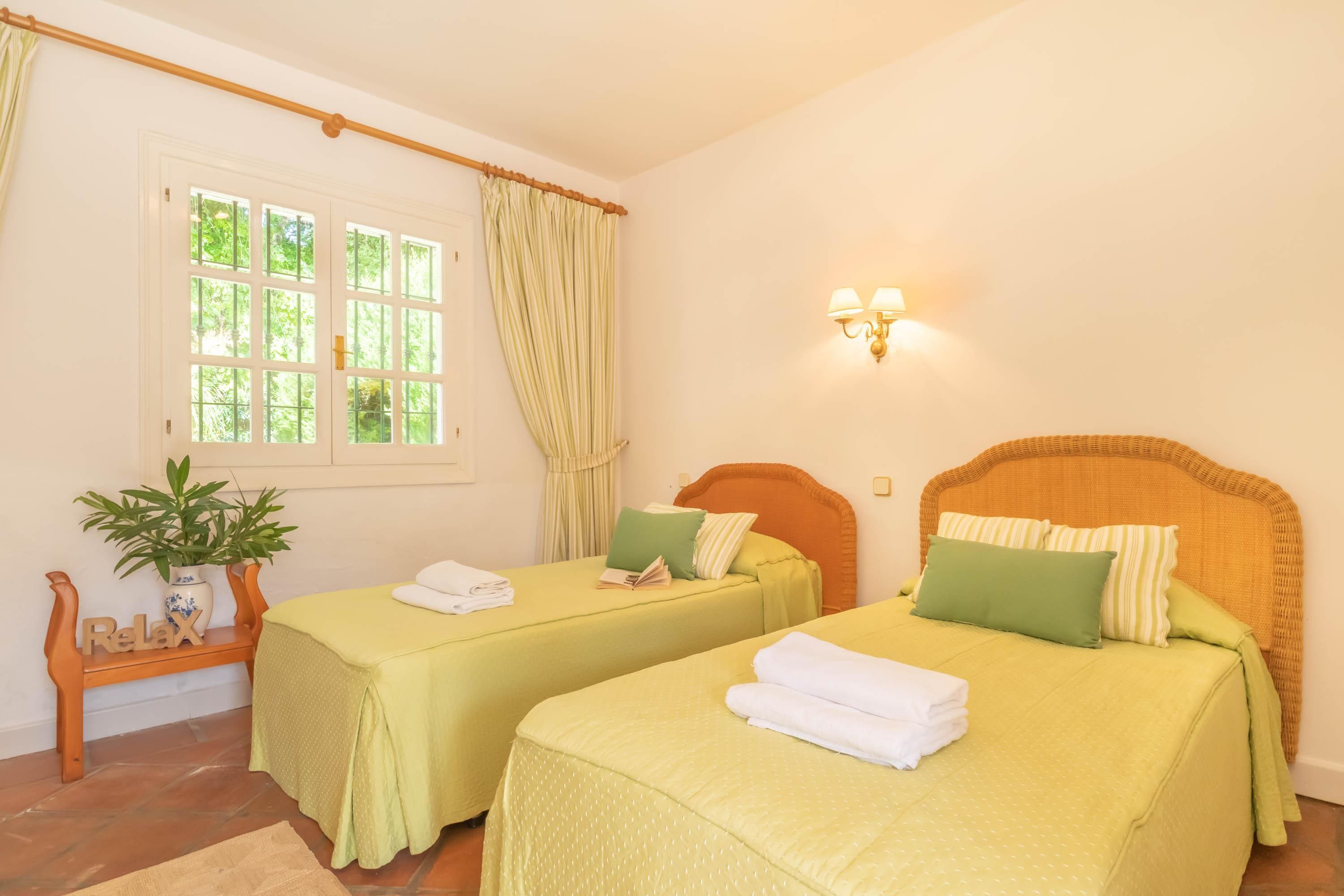 A room with two beds, a window with curtains, a bedside table with a plant, and a wall-mounted light fixture.