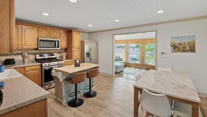A modern kitchen with wooden cabinets, stainless steel appliances, a central island with bar stools, and a dining area with a marble table.