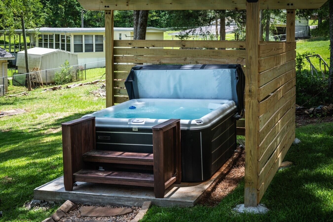 Un jacuzzi bajo una pérgola de madera en un patio trasero con césped y una valla al fondo.