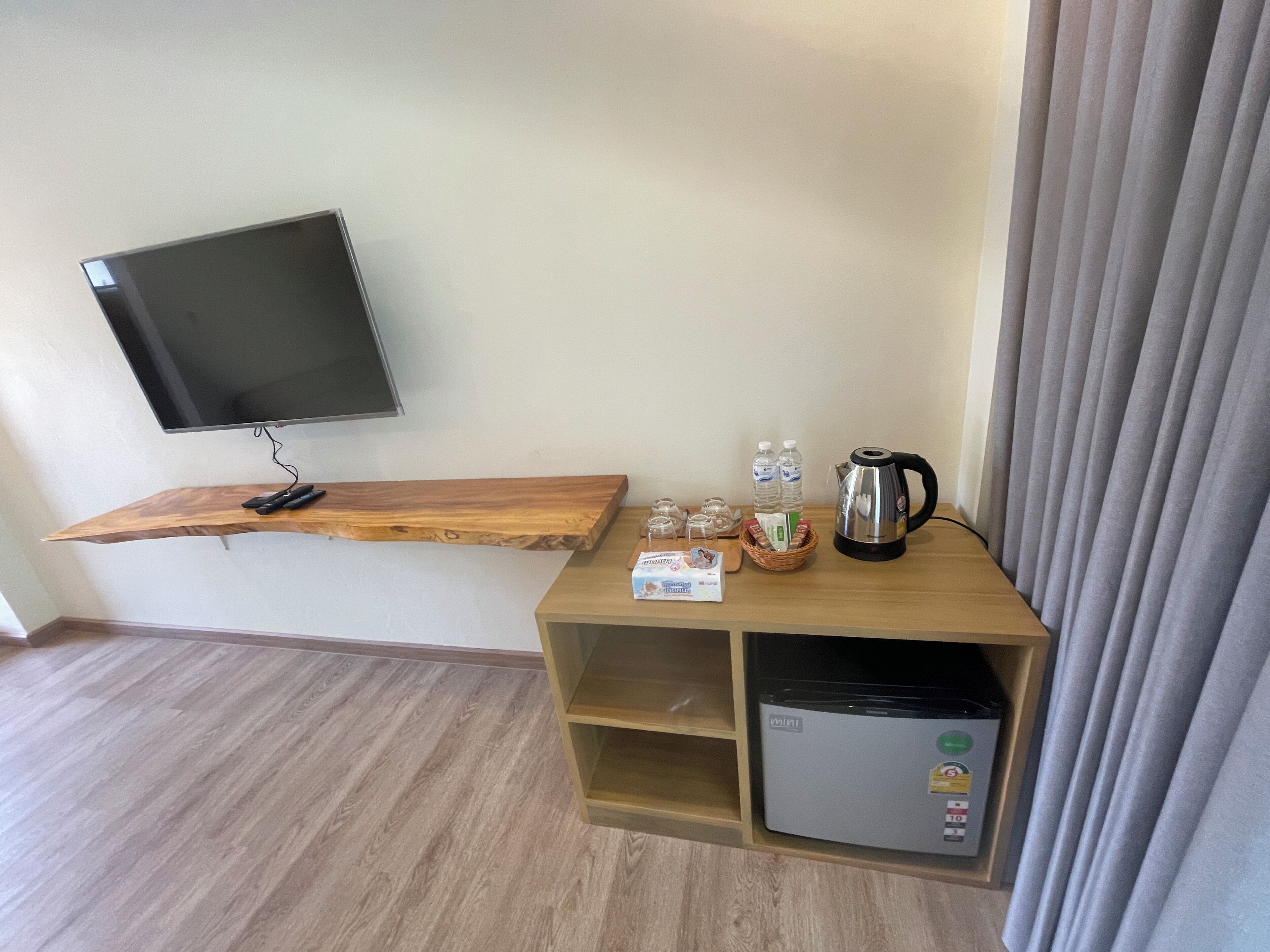 A hotel room with a mounted TV, wooden shelf with remote controls, a mini fridge, a kettle, and a basket with bottled water and snacks.