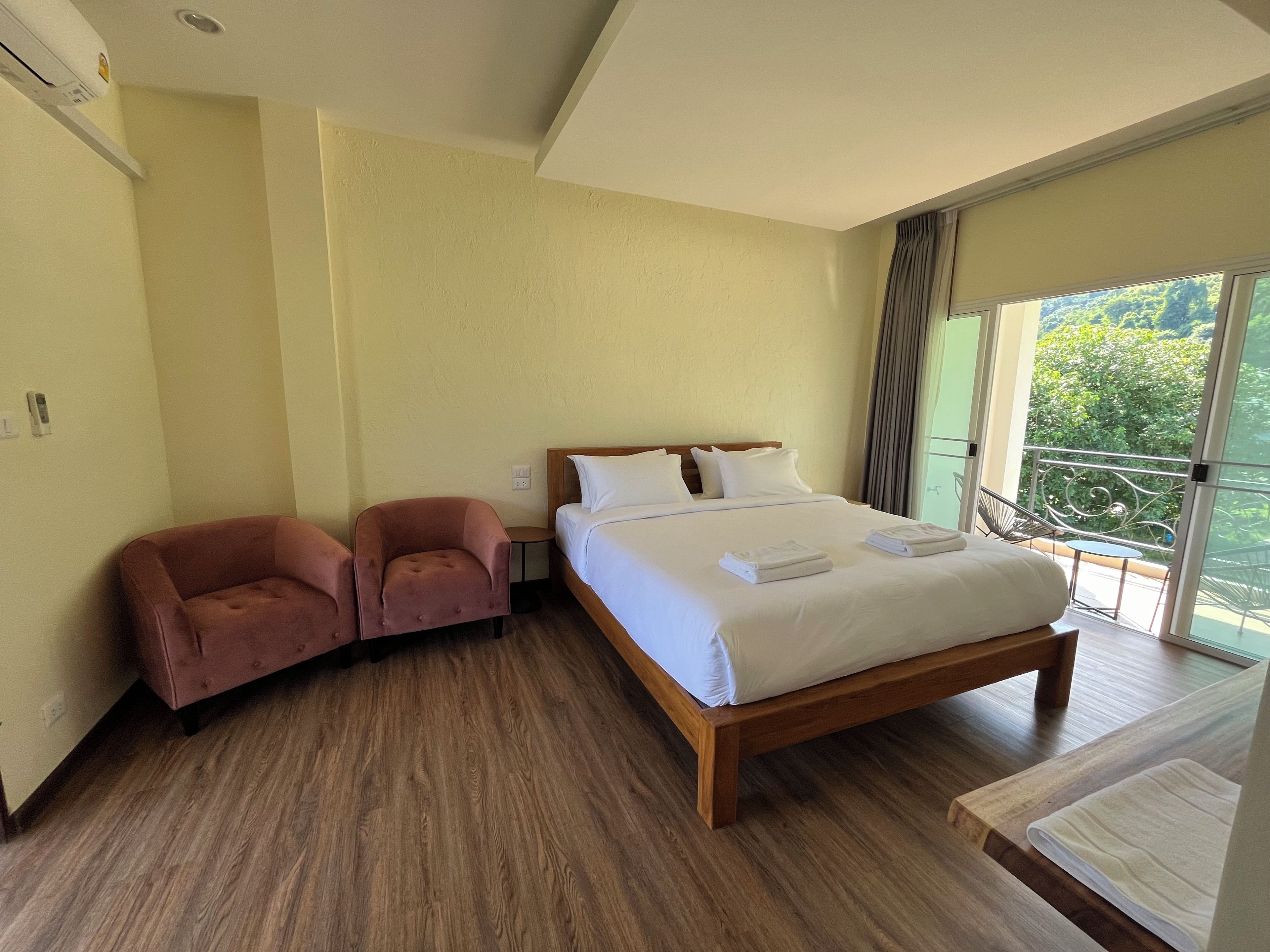 A hotel room with a large bed, two armchairs, a small table, and a balcony with a view of greenery.