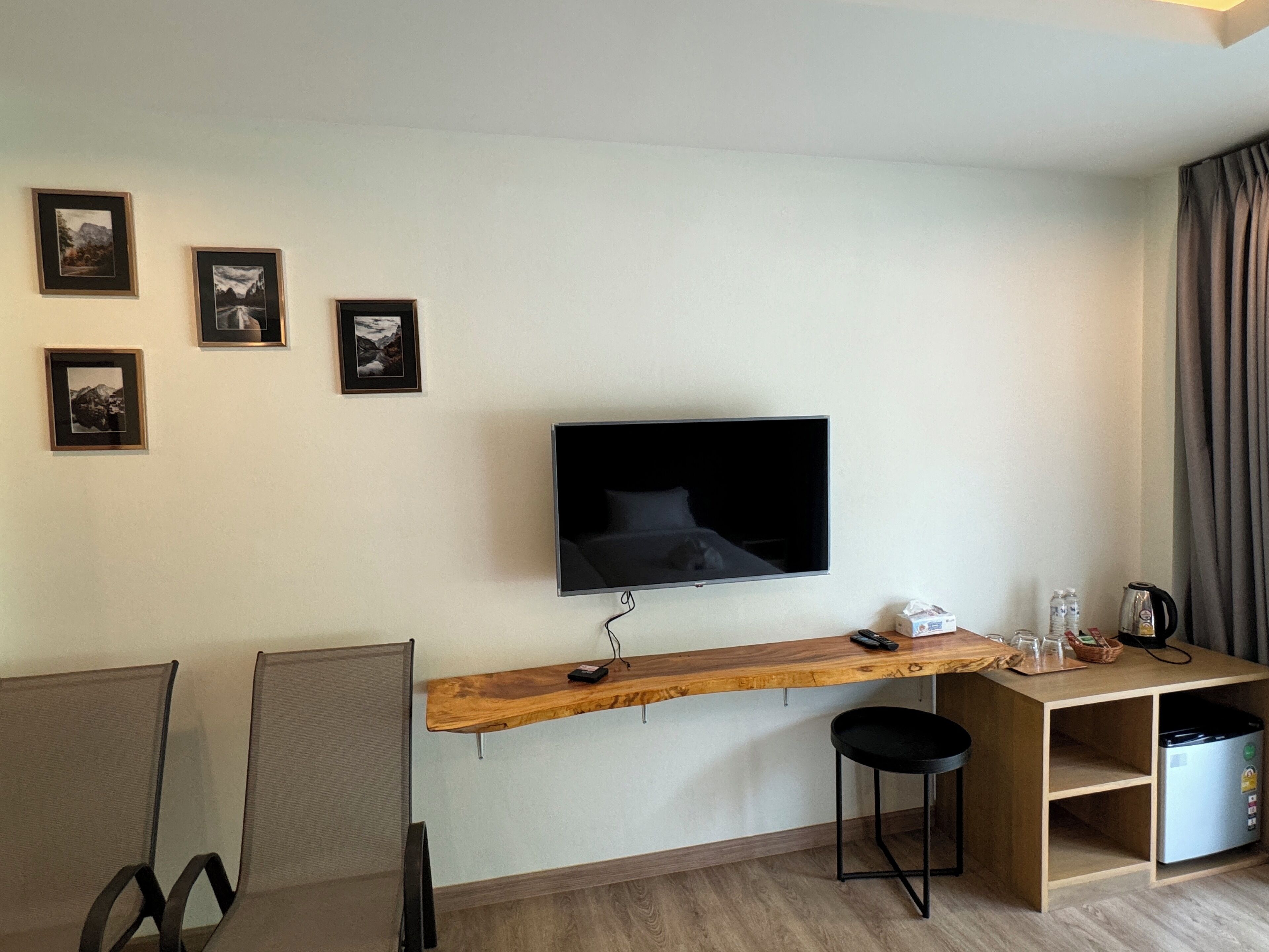 A modern room with a mounted TV, a wooden desk, a chair, a small stool, and a shelf with a kettle and glasses.