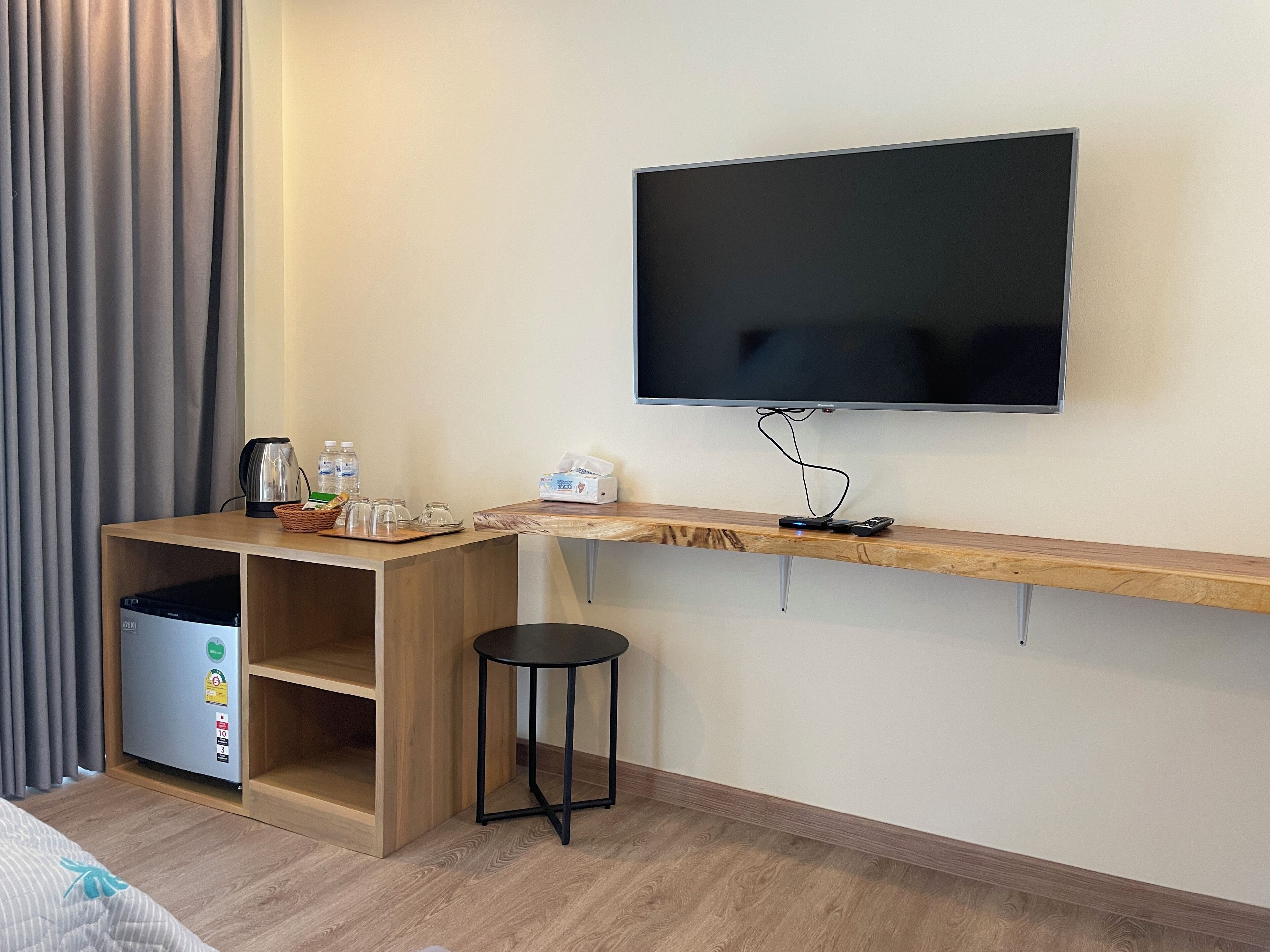 A hotel room with a wooden desk, a flat-screen TV mounted on the wall, a small round table, and a mini-fridge.