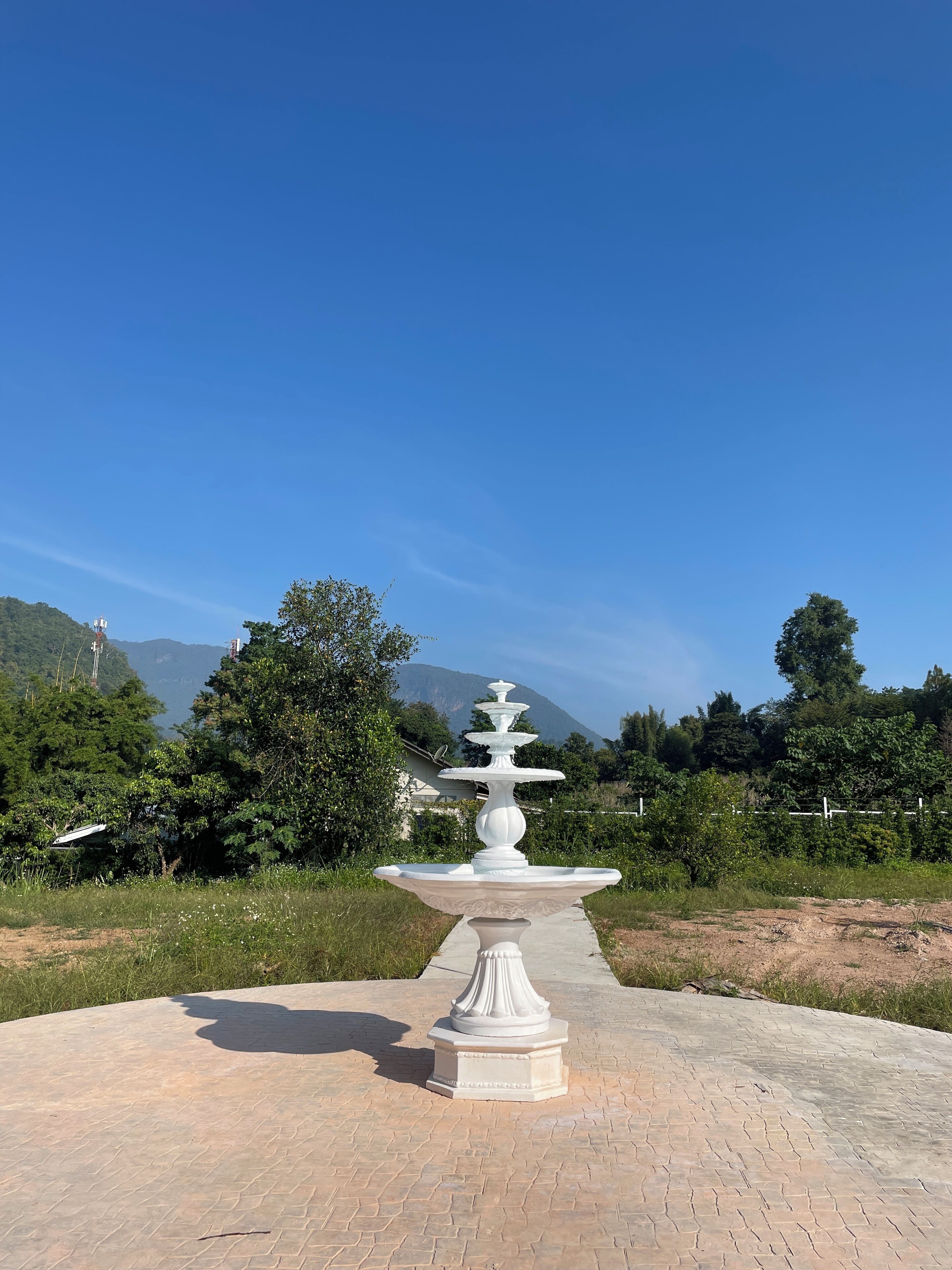 Una fuente blanca al aire libre con diseño escalonado, ubicada en un área pavimentada con un cielo azul despejado y montañas al fondo.