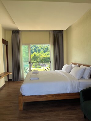Habitación de hotel con una cama grande, cabecera de madera, un sillón verde y un balcón con vistas a zonas verdes.