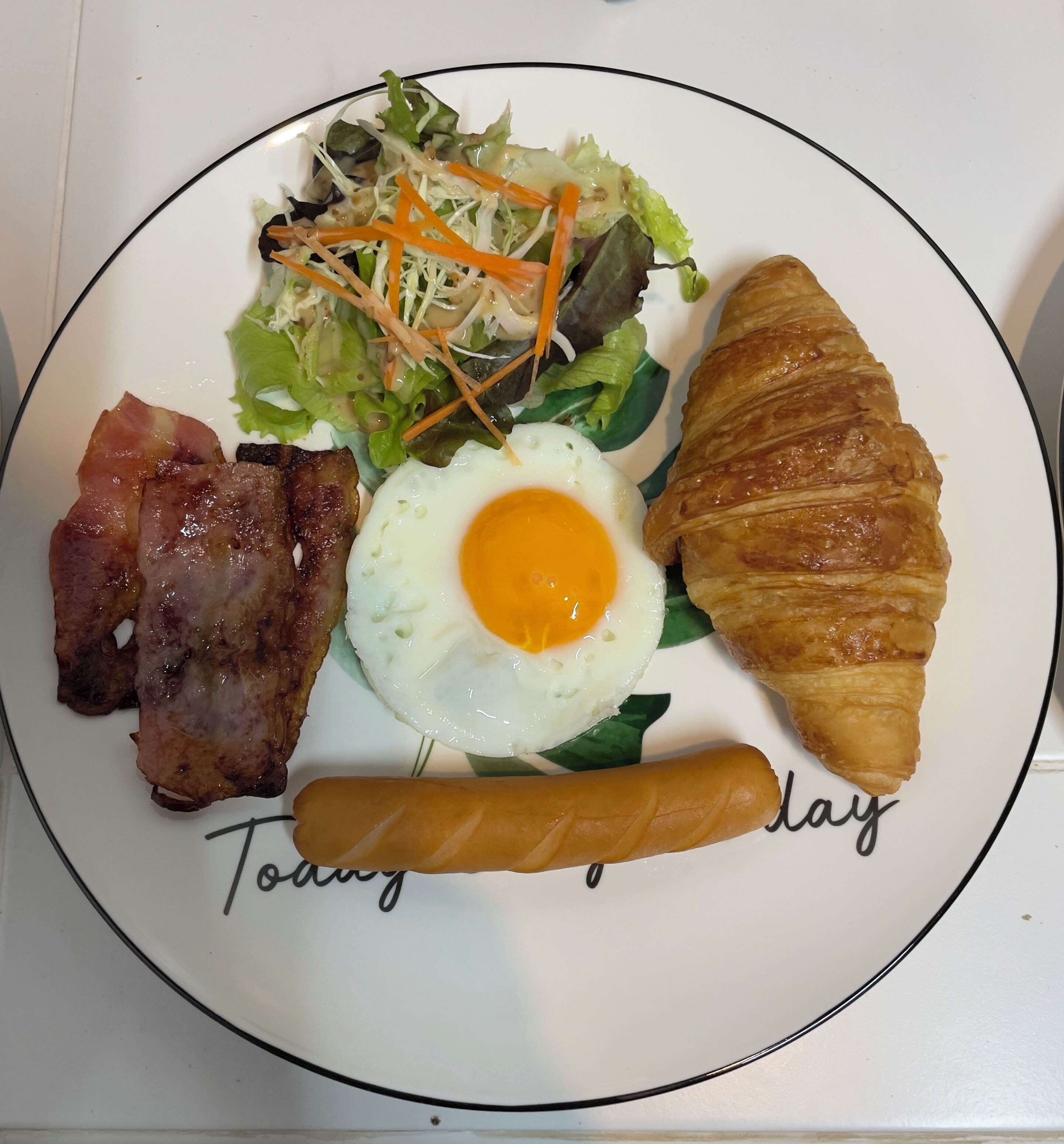 Un plato con un croissant, tocino, un huevo frito, salchicha y una ensalada.
