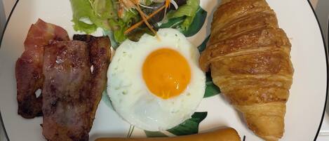 Un plato con un croissant, tocino, un huevo frito, salchicha y una ensalada.