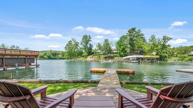 Terraza junto al lago con dos sillas Adirondack y una mesa de picnic, con vista a un lago tranquilo y un muelle.