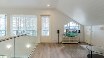 Una habitación amplia con suelo de madera, barandilla de cristal y un gran ventanal.