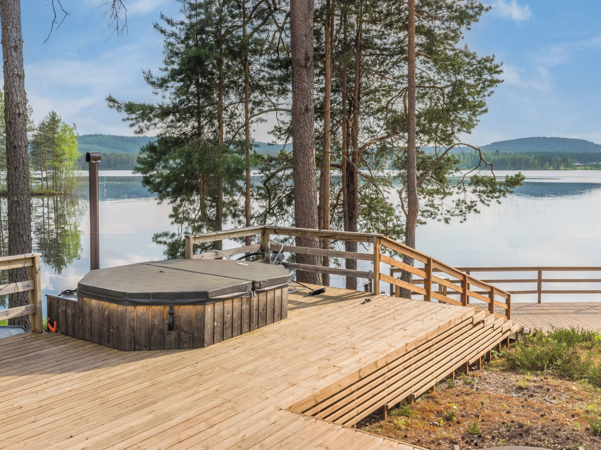 Un terrazza in legno con vasca idromassaggio, circondata da alberi e affacciata su un lago tranquillo.