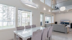 Un comedor moderno con una mesa de vidrio y sillas grises, una cocina con televisor y ventanas grandes con persianas.
