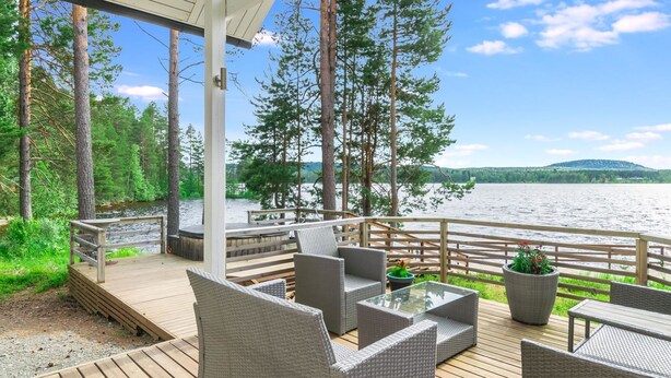 Una terraza techada con mobiliario de mimbre, una mesa de vidrio y vistas a un lago y un bosque.