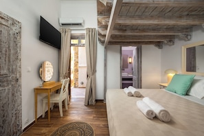 A bedroom with a wooden desk, a bed with rolled towels, a TV, and a bathroom visible through an open door.