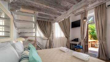 A hotel room with a bed, a TV, a window with curtains, a small table with a lamp, and a view of greenery outside.