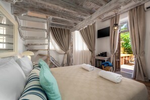 A hotel room with a bed, a TV, a window with curtains, a small table with a lamp, and a view of greenery outside.
