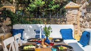 A dining area with a wooden table set for a meal, a sofa with blue cushions, and a stone wall in the background. - 4U Rhodes (Rhodes)