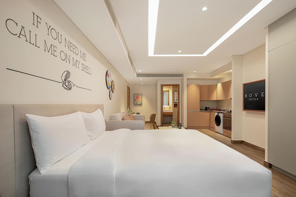 A modern hotel room with a large bed, a small seating area, a kitchenette, and a bathroom.