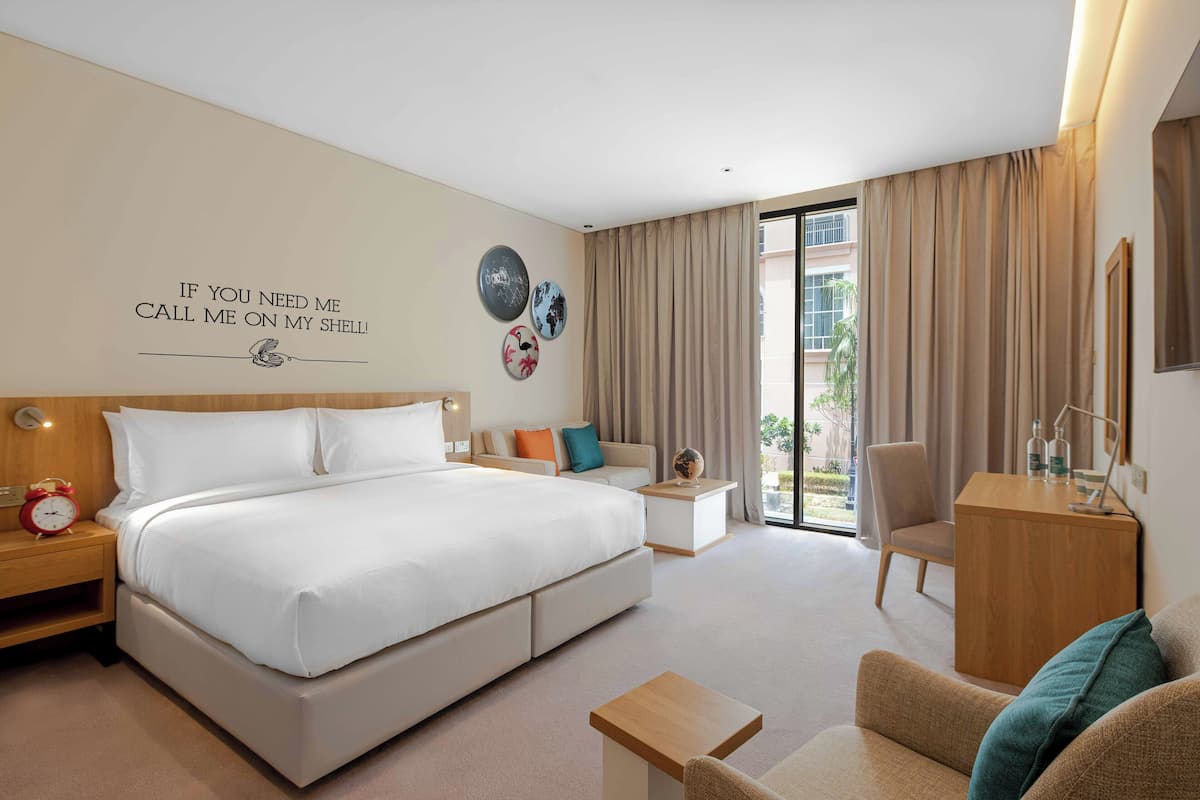 A modern hotel room with a large bed, a sofa, a desk, and a view of the outdoors.
