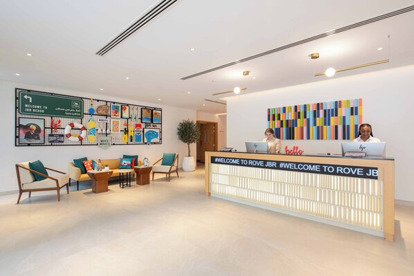 Un'area reception con scrivania, due sedie, un divano e una parete decorata con manifesti e un cartello di benvenuto per gli ospiti a JBR Beach.