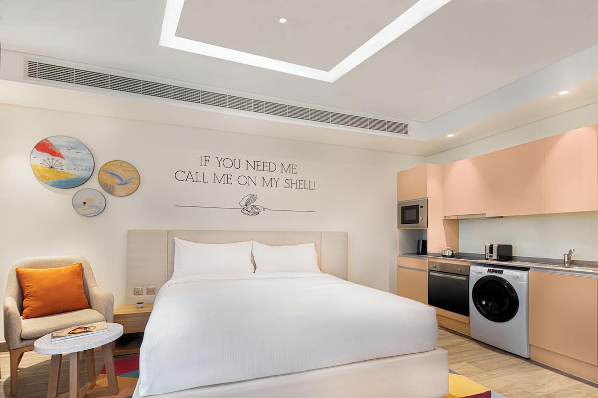 A modern hotel room with a bed, a chair, a small table, a washing machine, and a kitchen area.