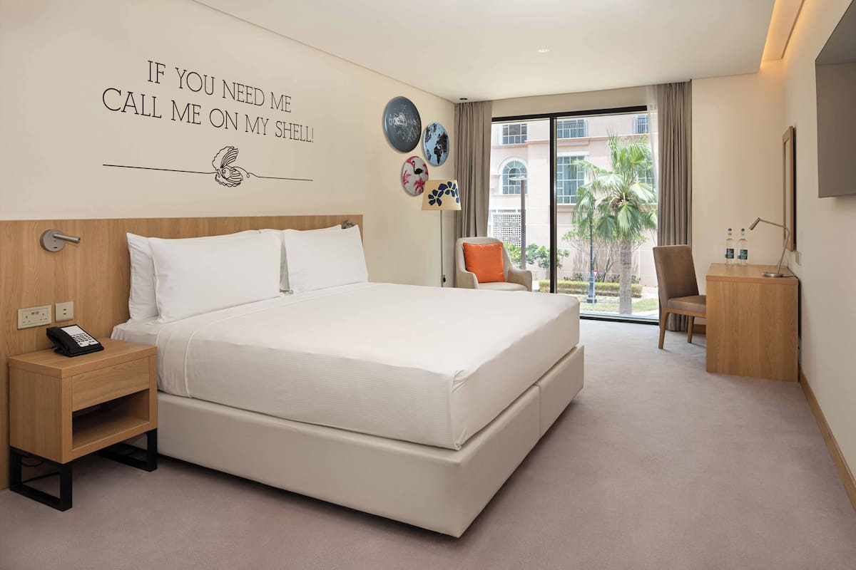 A modern hotel room with a large bed, a bedside phone, a desk with a lamp, and a chair.