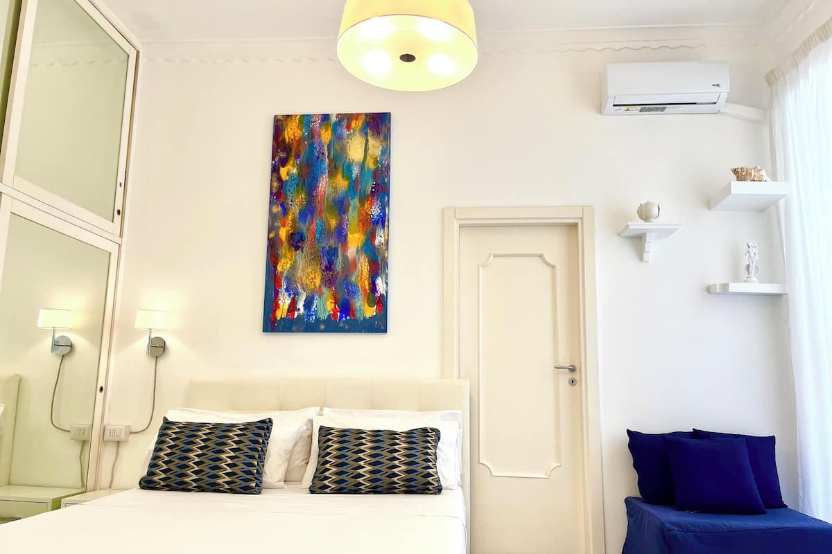 A hotel room with a bed, bedside lamps, a wall-mounted air conditioner, a door, and a shelf with decorative items.