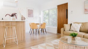 A modern living room with a sofa, a round glass coffee table, a dining area with chairs, and a wooden door.