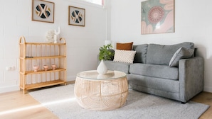 A living room with a grey sofa, a round wicker coffee table, a wicker shelf with decorative items, and framed artwork on the walls. - The Byron - Luxury Seaside Kingston Park Studio (Kingston Park)