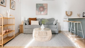 A living room with a grey sofa, a round wicker coffee table, a white vase, a wooden side table, and a white wall with artwork. - The Byron - Luxury Seaside Kingston Park Studio (Kingston Park)