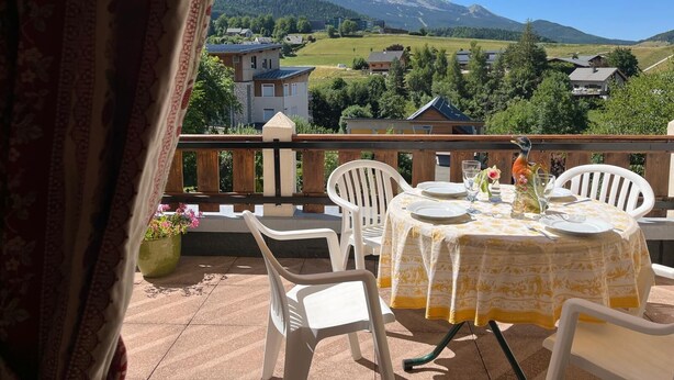 A balcony with a table set for two, overlooking a mountainous landscape with houses.