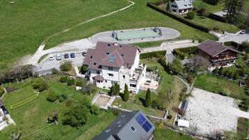 Une grande maison à deux étages avec un court de tennis, entourée de verdure et dotée d’une allée où sont garées des voitures.