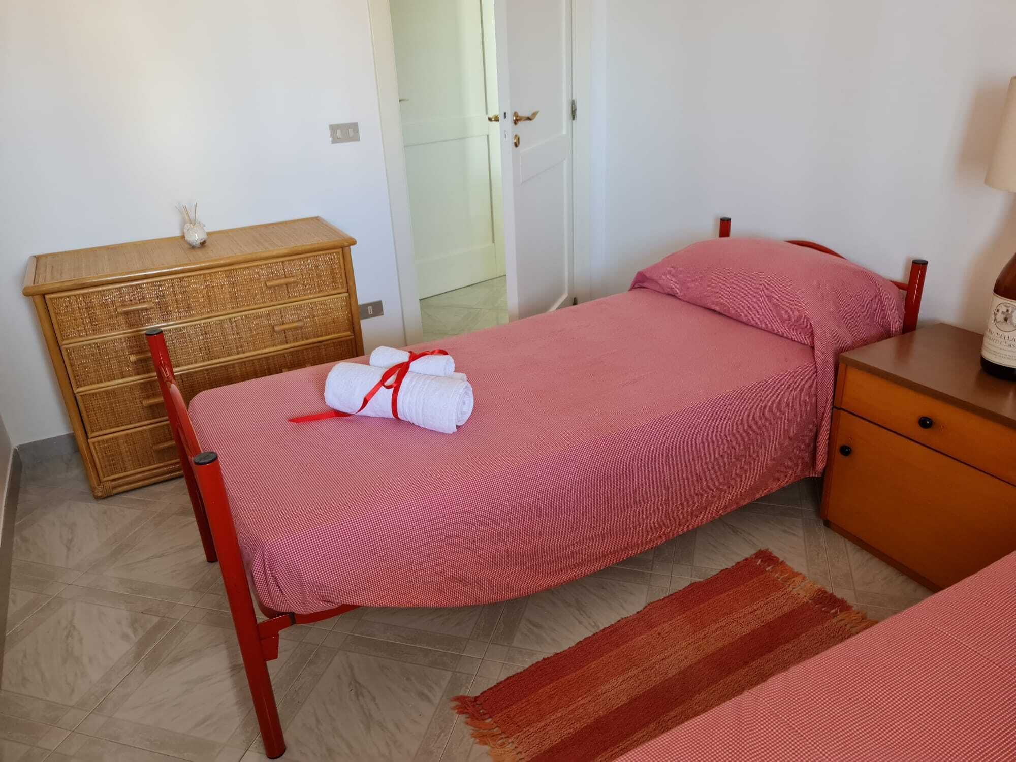 Un lit simple avec une couverture et des oreillers rouges, une commode en bois, une chaise rouge et une petite table sur laquelle se trouve une bouteille.