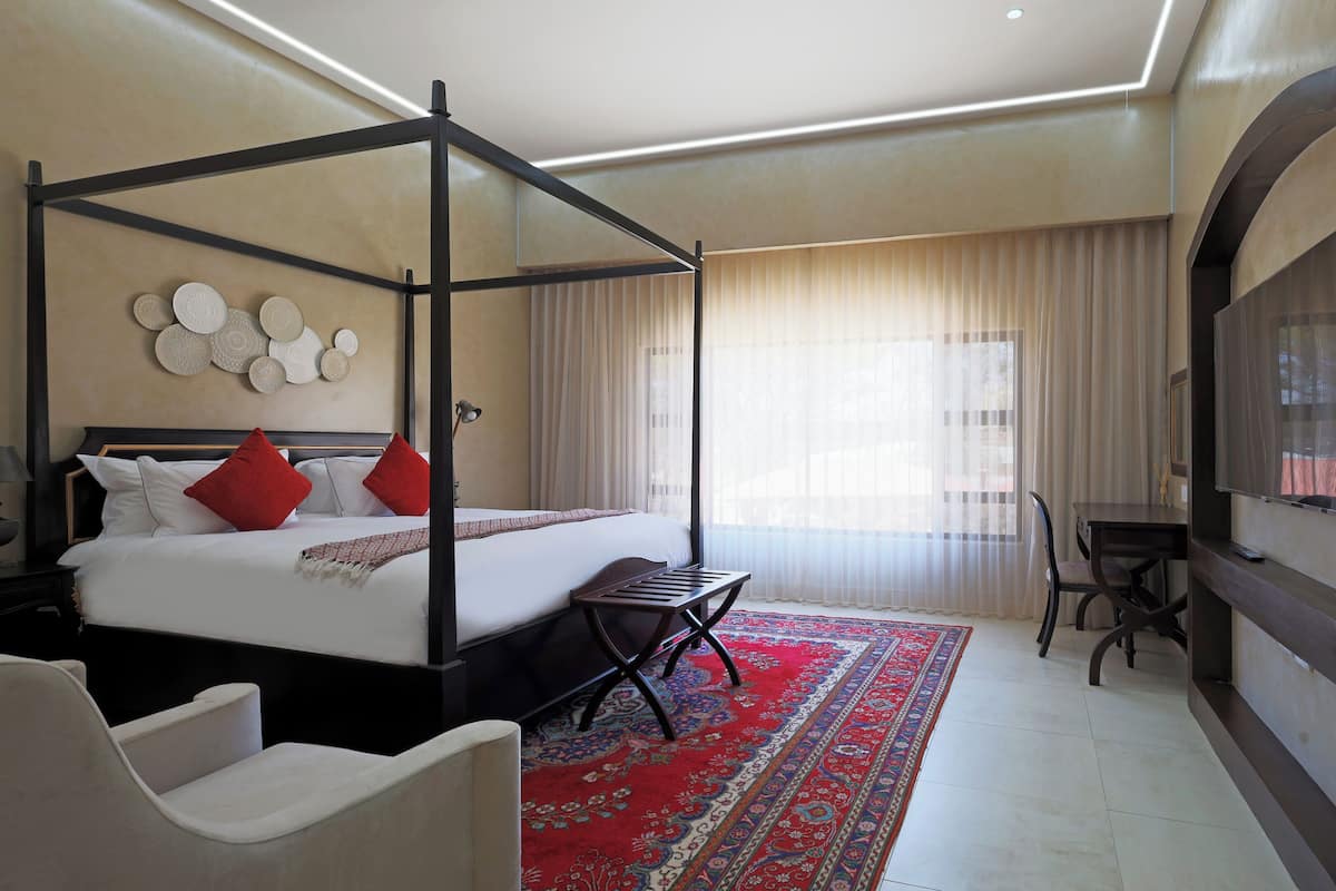 Ein Hotelzimmer mit einem Himmelbett, einem Teppich mit blau-rotem Muster, einem Schreibtisch mit Stuhl und einem großen Fernseher an der Wand.