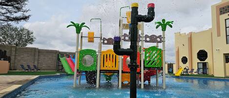 Un espace de jeux aquatiques avec des toboggans et une piscine, entouré d’un mur de briques et d’un bâtiment doté de fenêtres.