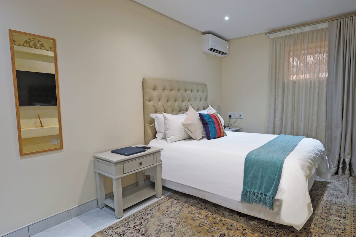 Ein Hotelzimmer mit einem großen Bett, einem Nachttisch, einem Spiegel, einer Klimaanlage, einem Fenster mit Vorhängen und einem gemusterten Teppich.