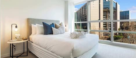 A modern hotel room with a large bed, a bedside table with a lamp, and a view of a cityscape through large windows.