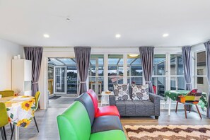 A modern living room with a sofa, a dining table, and a view through large windows. - Ellerslie 3BR House | Fresh Reno | Zodiak Stays (Auckland)