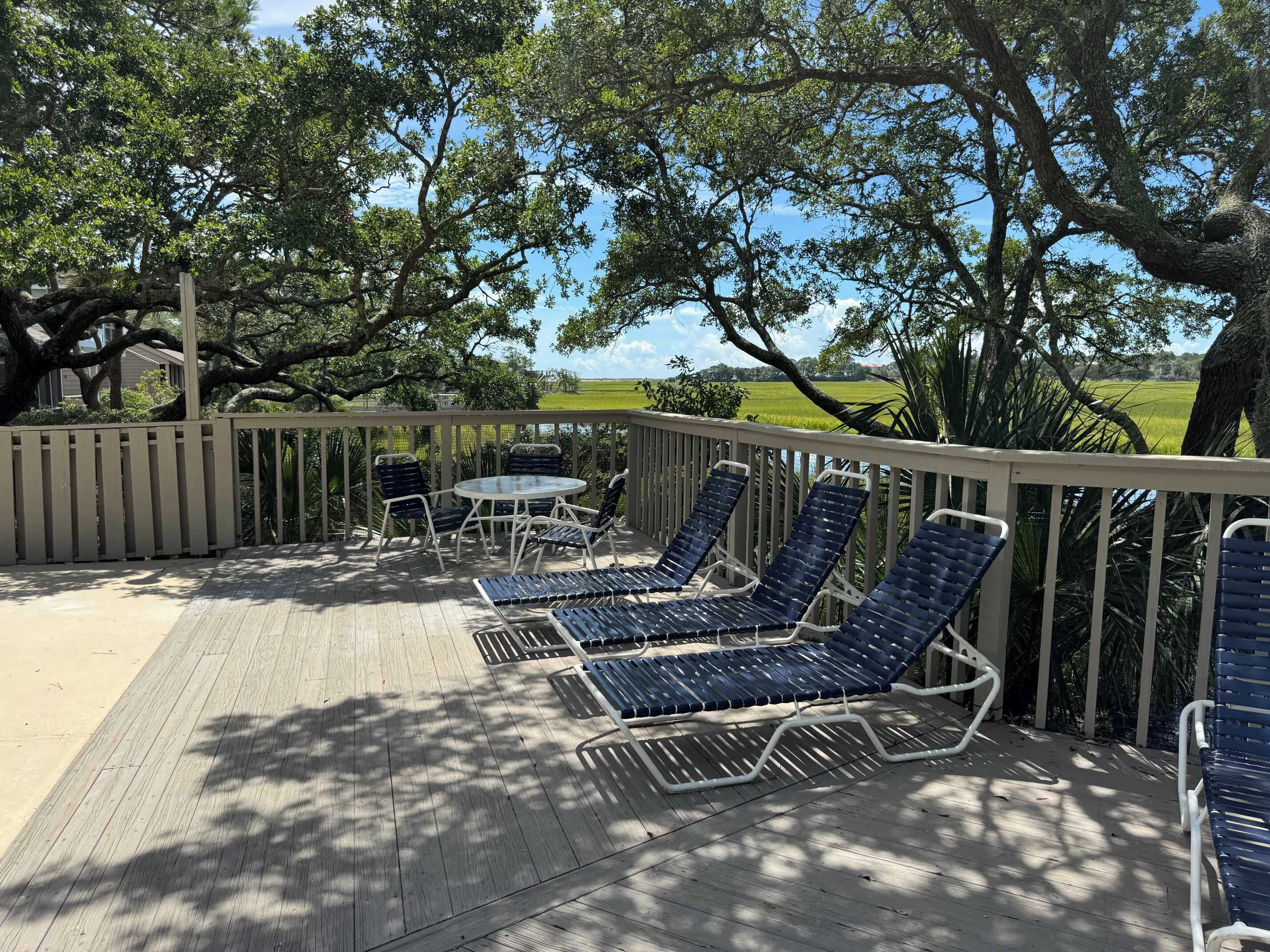 A covered outdoor area with a wooden deck, lounge chairs, and a view of a green field and water.