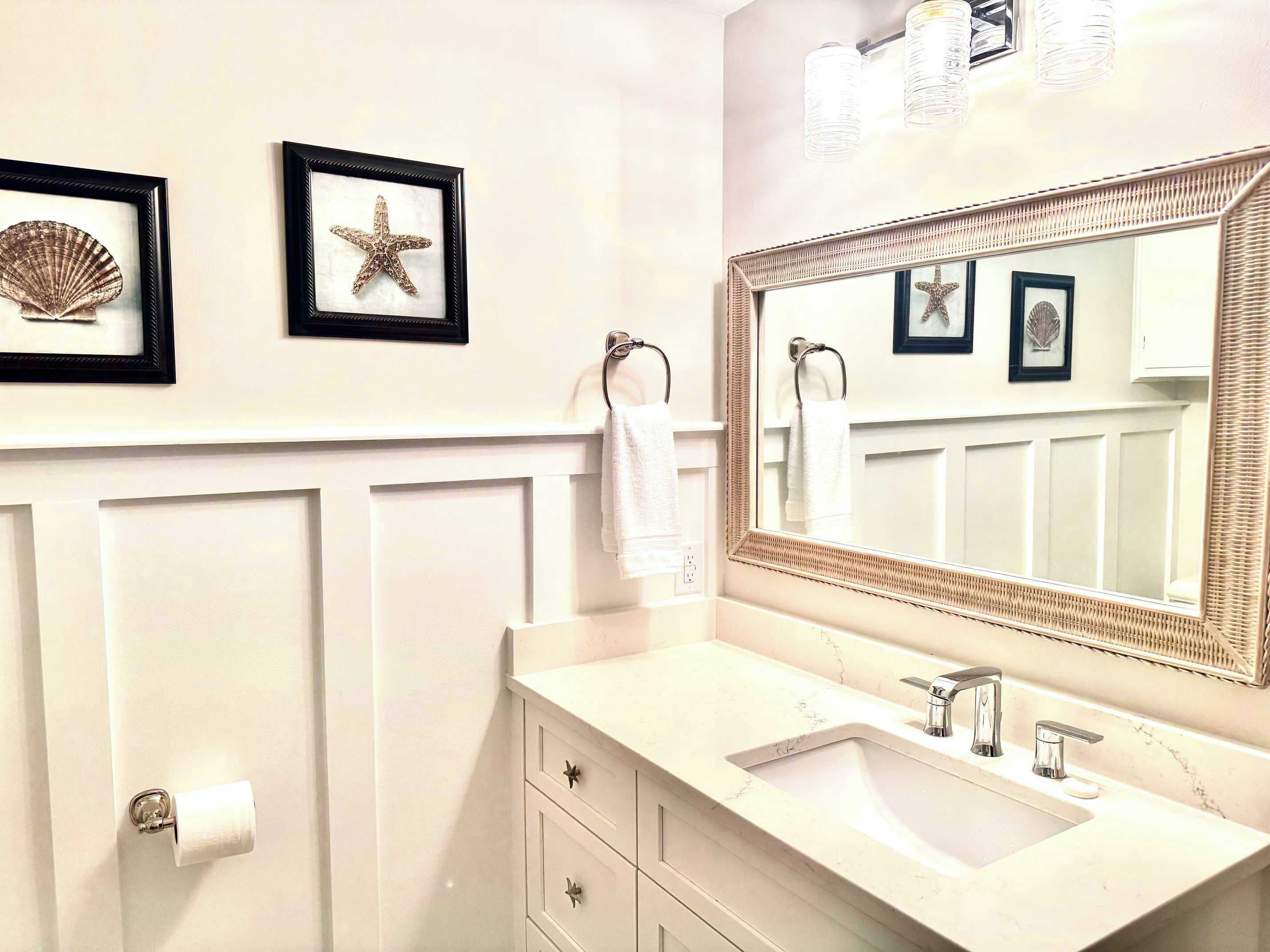 A bathroom with a white vanity, a large mirror, and framed sea shell and starfish decorations on the wall.