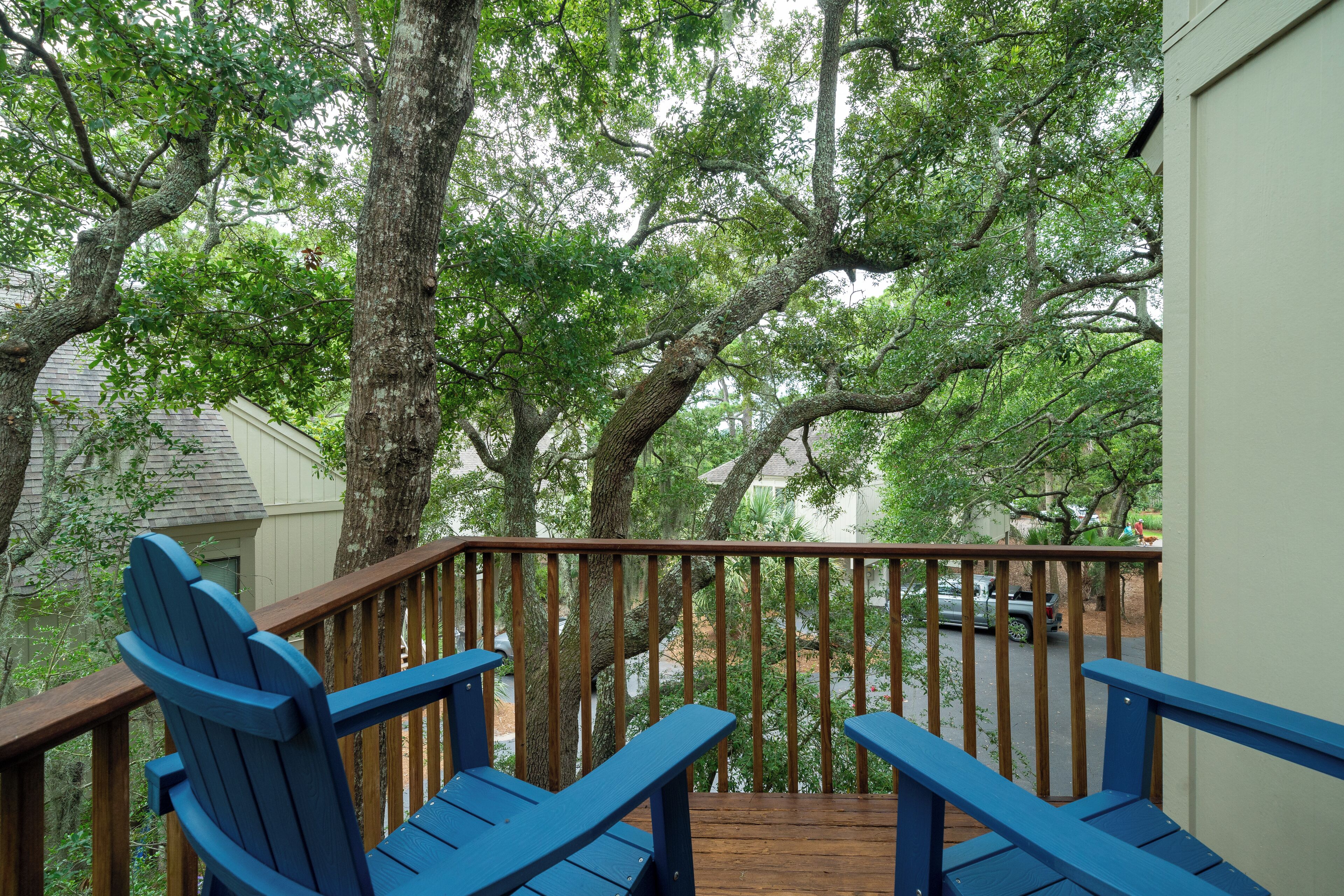 A wooden deck with blue Adirondack chairs, surrounded by trees and a view of a street.