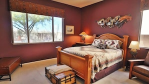 Quarto com uma janela grande, uma cama de madeira, um criado-mudo, um banco e um objeto decorativo na parede.