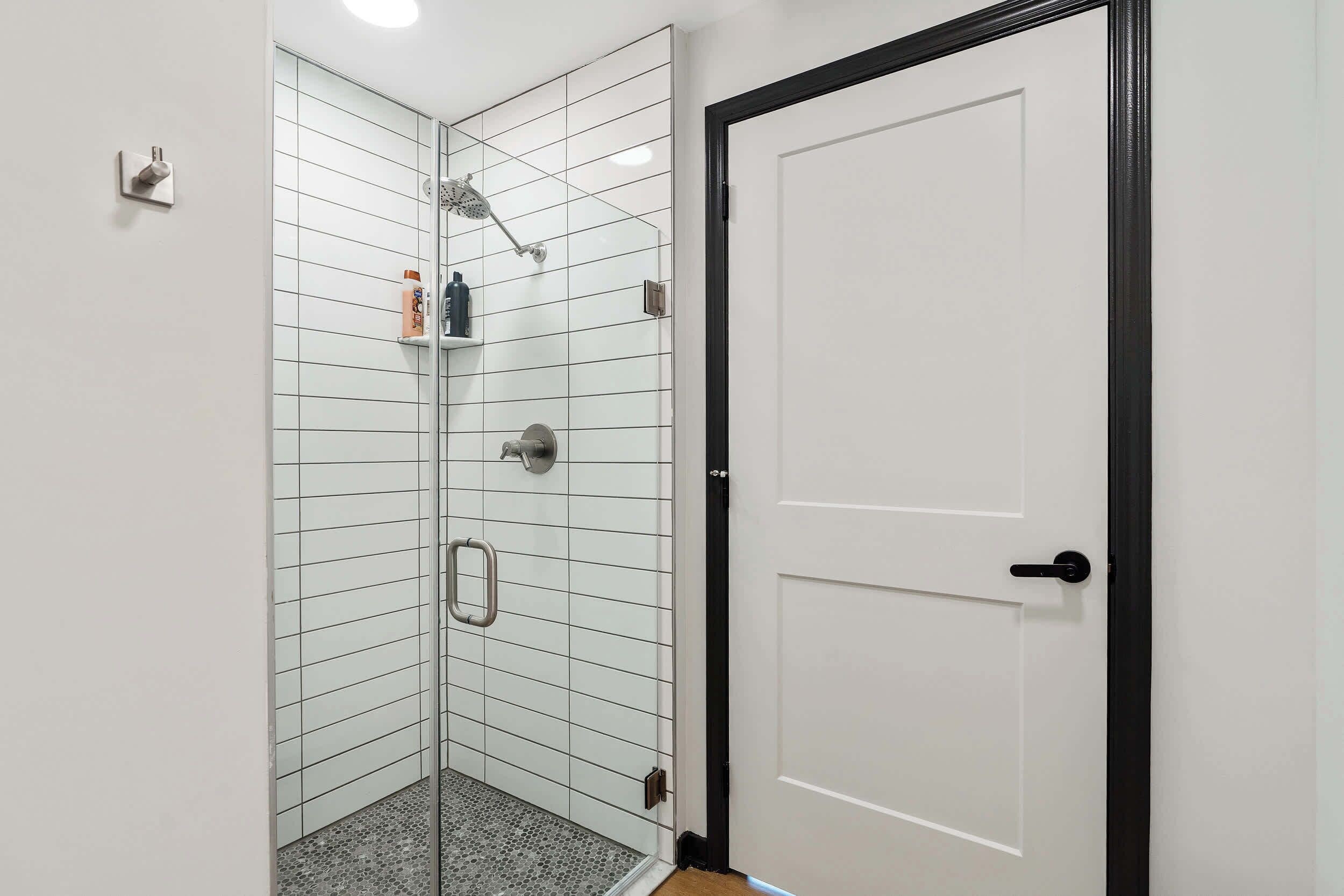 Ducha en esquina con mampara de vidrio, pared de azulejos blancos y puerta blanca con marco negro.