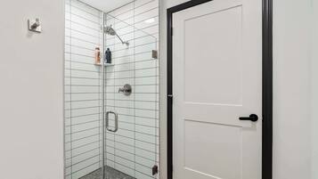 Ducha en esquina con mampara de vidrio, pared de azulejos blancos y puerta blanca con marco negro.