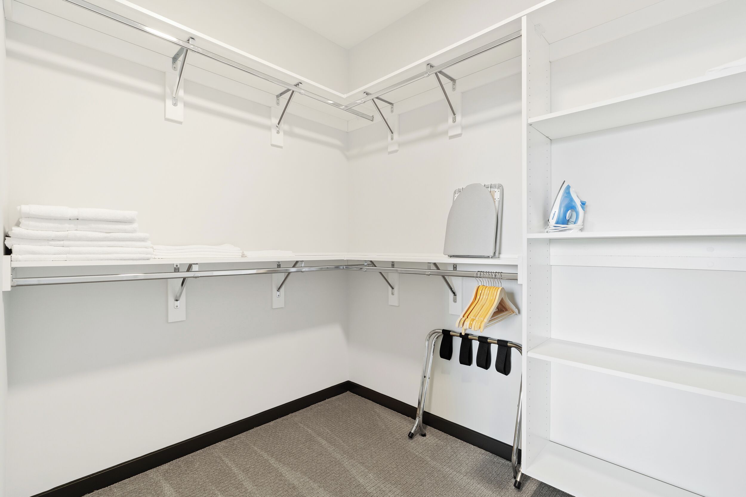 A hotel room with a white wall, a grey carpet, a metal rack with towels, a small ironing board, and a shelf with a blue object.