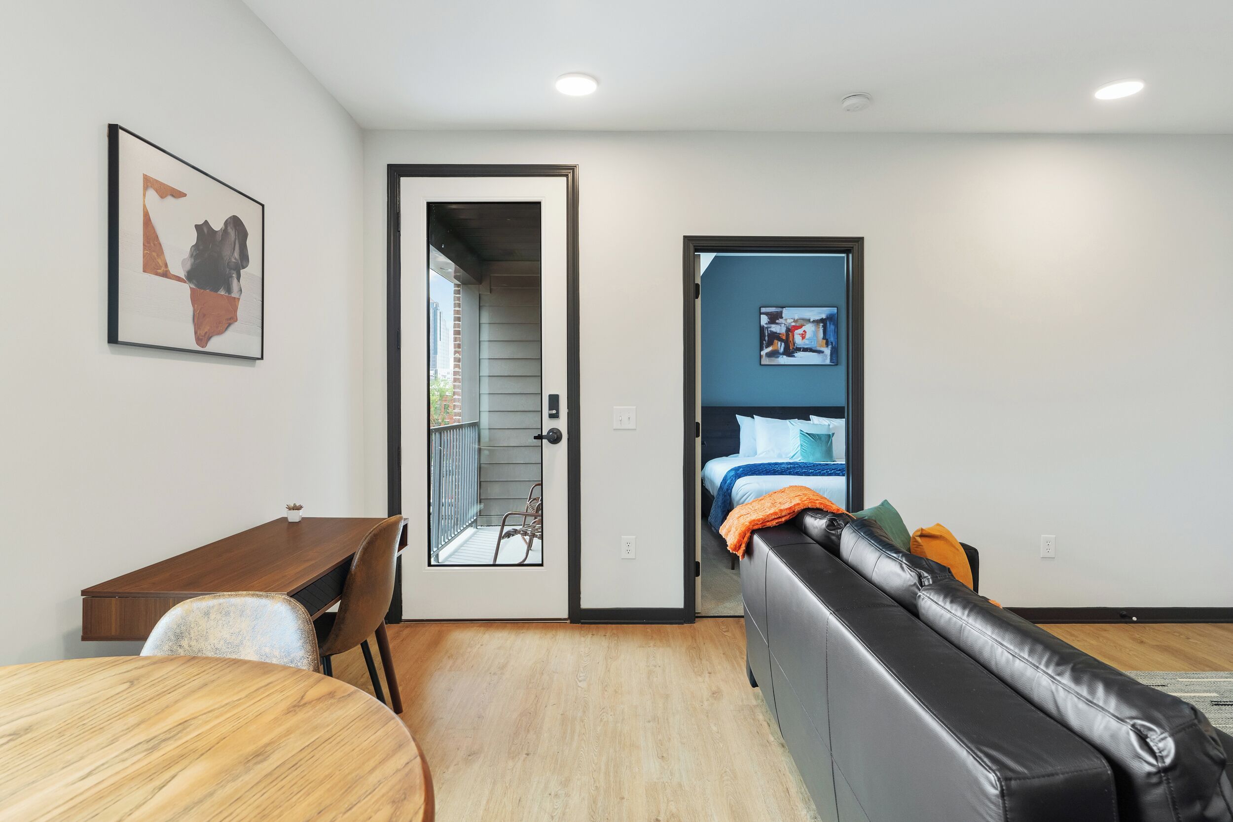 Une chambre moderne avec une table à manger en bois, un canapé en cuir noir, un lit avec une tête de lit bleue et une porte donnant sur un balcon.