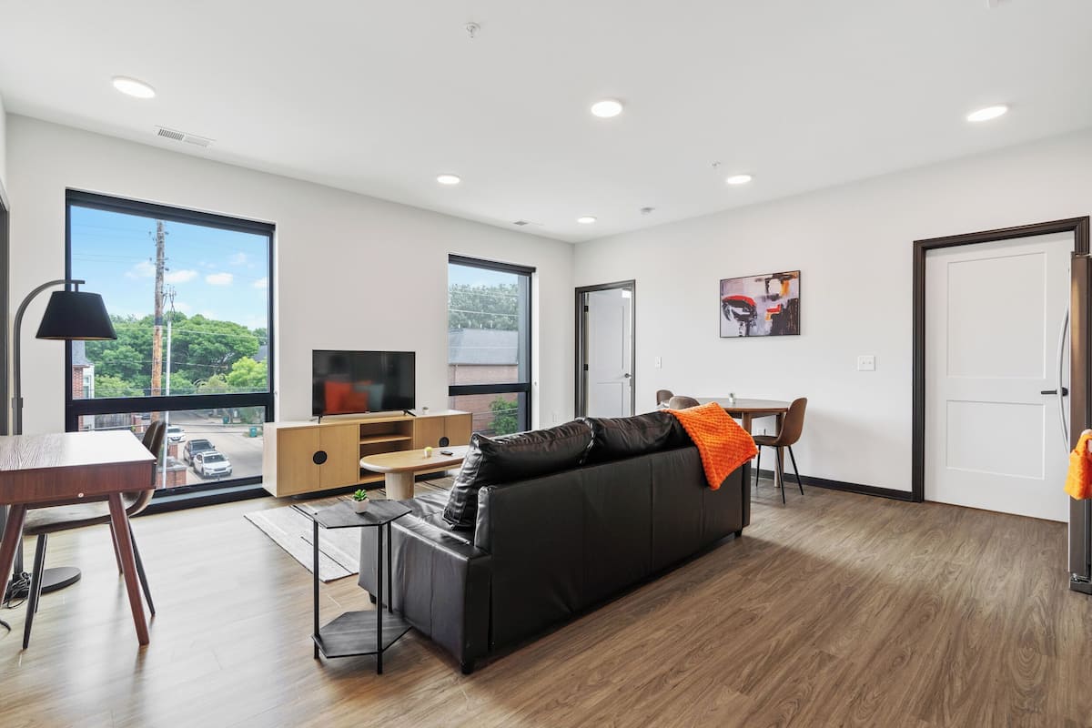 A modern living room with a black leather sofa, wooden flooring, a TV, and a dining area.