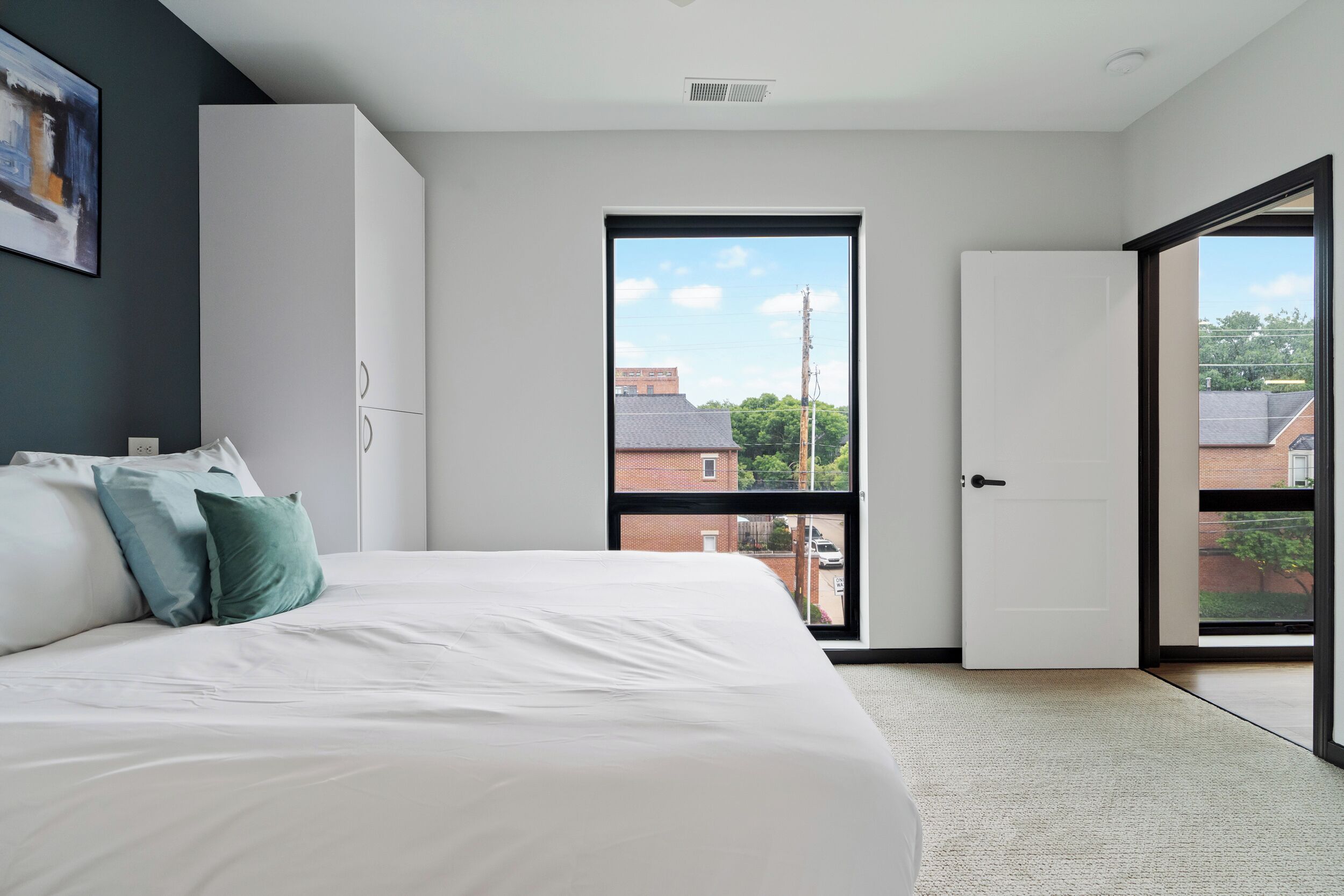 A bedroom with a large bed, a wardrobe, a door leading to a balcony, and a view of buildings and greenery outside.