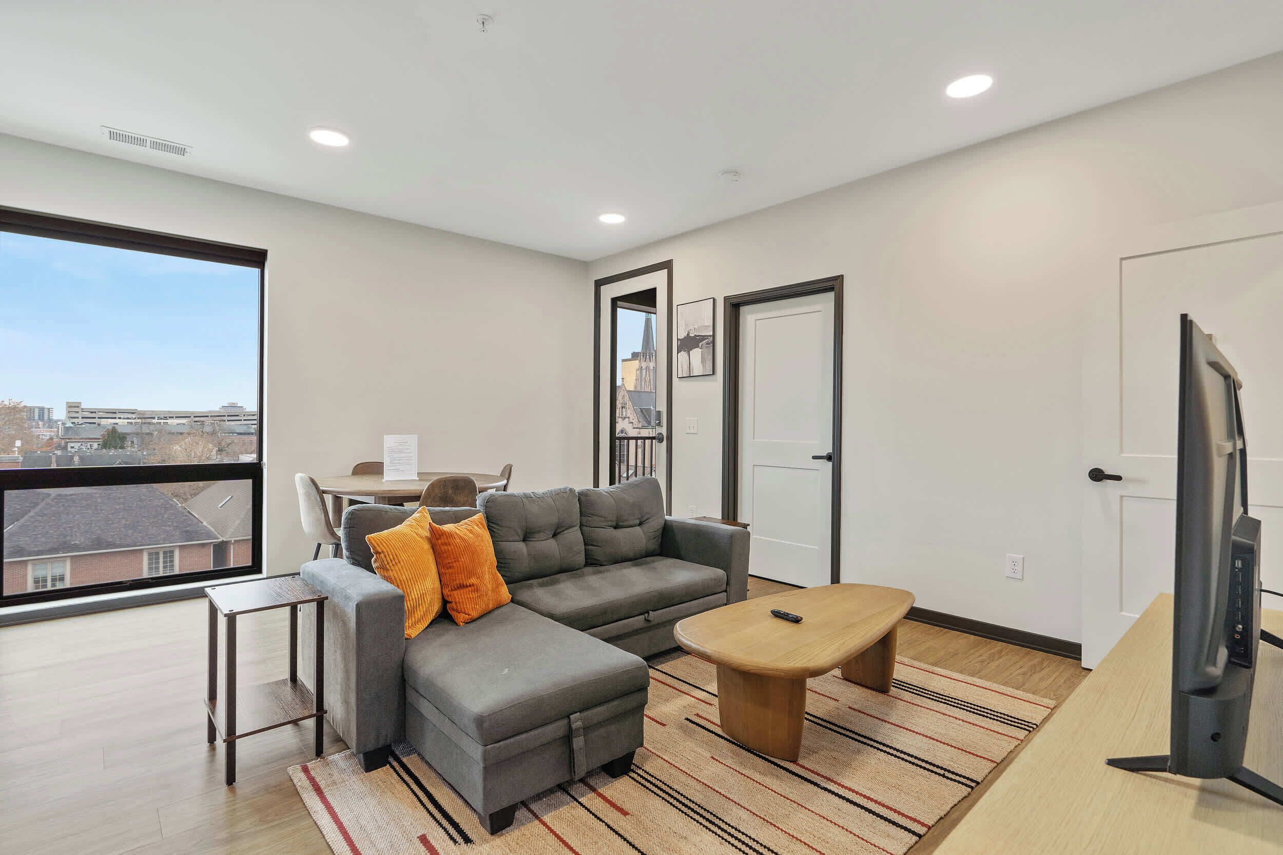 A modern living room with a grey sectional sofa, a small wooden coffee table, a TV, and a large window offering a city view.
