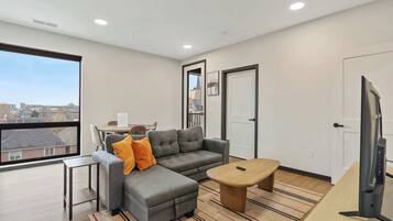 A modern living room with a grey sectional sofa, a small wooden coffee table, a TV, and a large window offering a city view.
