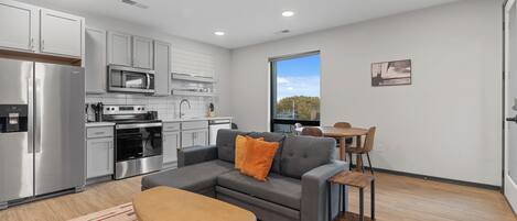 A modern kitchen with stainless steel appliances, a gray sofa with orange cushions, a wooden coffee table, and a dining area with a wooden table and chairs.